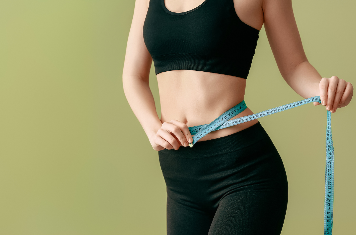 Woman in black athletic wear uses a blue measuring tape to measure her waist, standing against a green background, fitness-focused image.