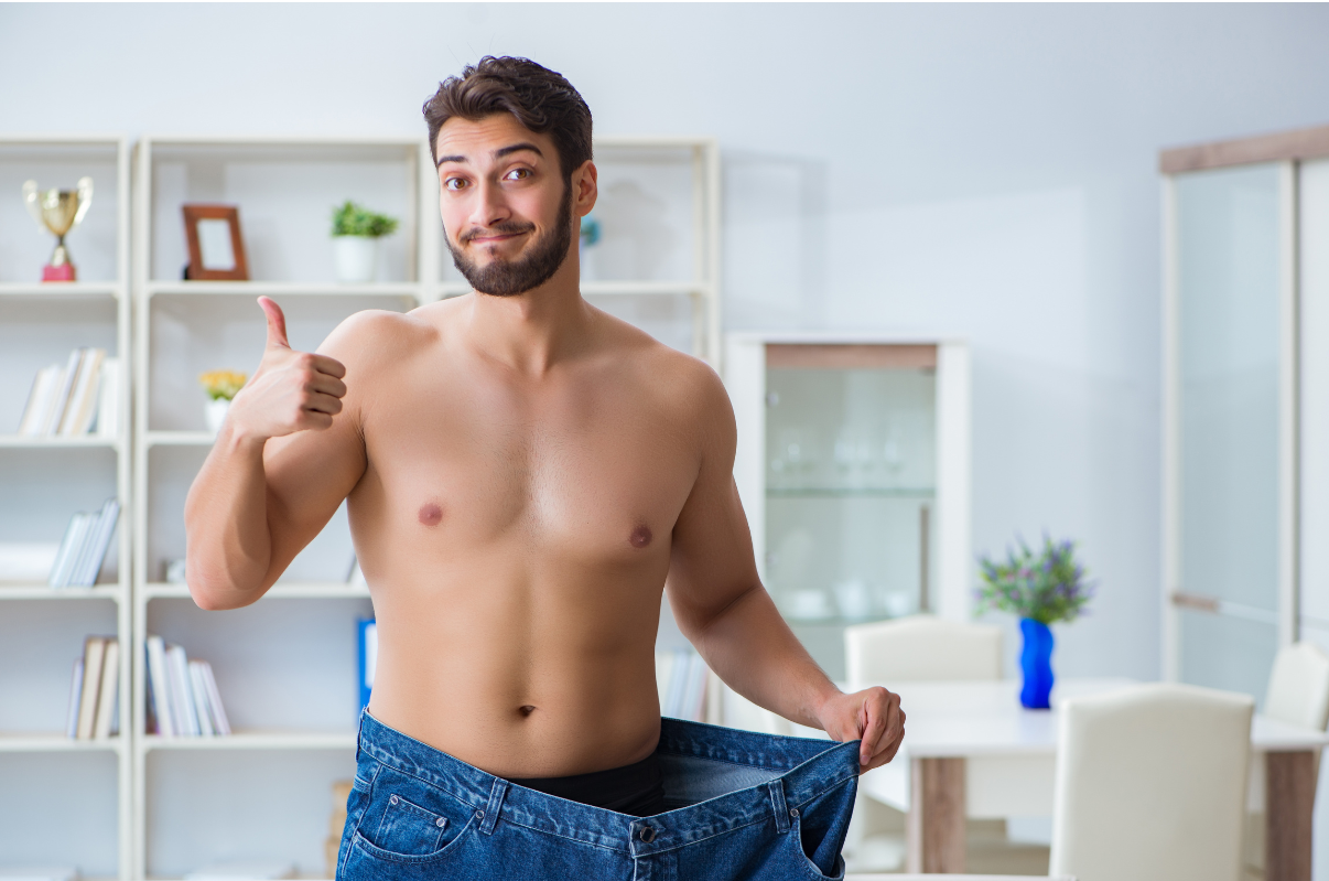 Smiling shirtless man in a bright living room holds out oversized jeans to show they no longer fit.