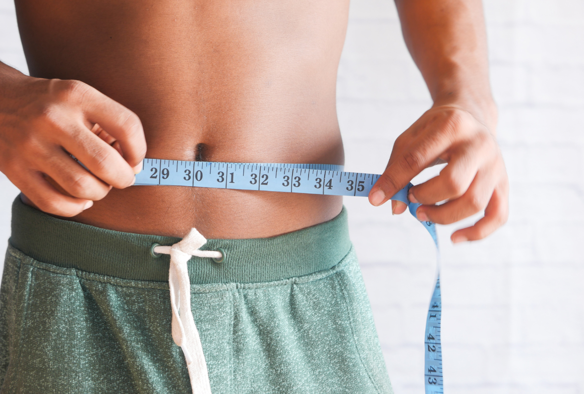 Person measures their waist with a blue measuring tape, wrapped around the abdomen over green shorts.