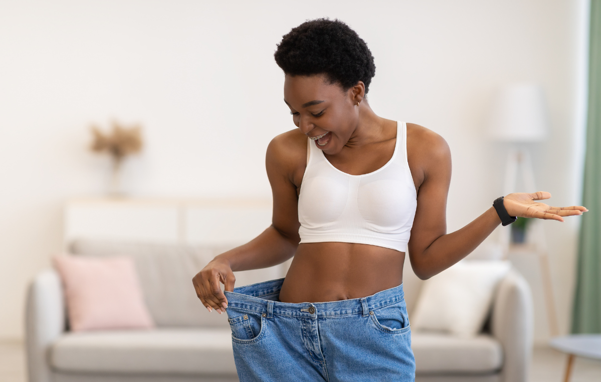 Woman in a white sports bra smiles as she checks that a pair of jeans is too large for her, indoors in a living room.
